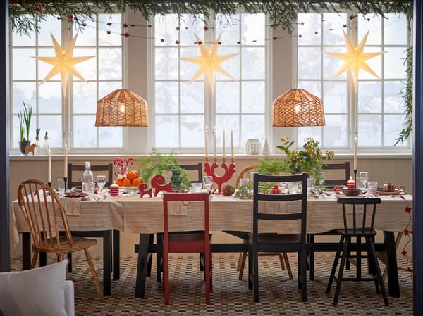Sala da pranzo con un grande tavolo apparecchiato per la cena di Natale con sedie di diverse forme e colori accanto a un albero di Natale, sopra il quale sono appese alcune luci natalizie.