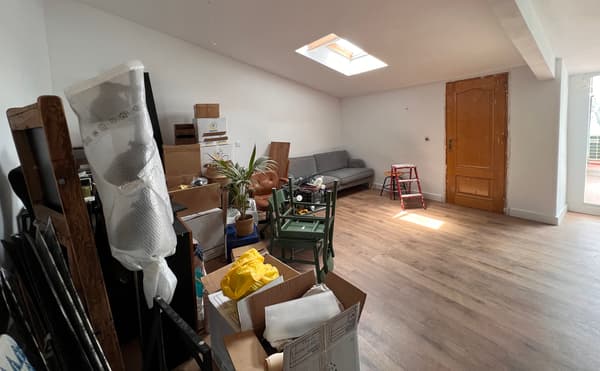 Sala d'un àtic petit abans de la reforma. Té un sofà, una escala i diversos productes en caixes i embalats.