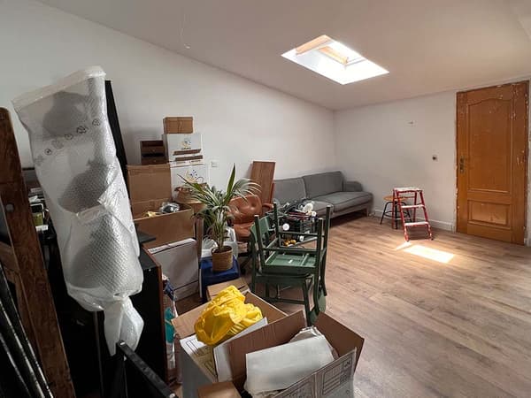 Sala sota teulada i amb una finestra al sostre abans de la reforma, amb mobles amuntegats en un costat i un sofà i una escala en l'altre.