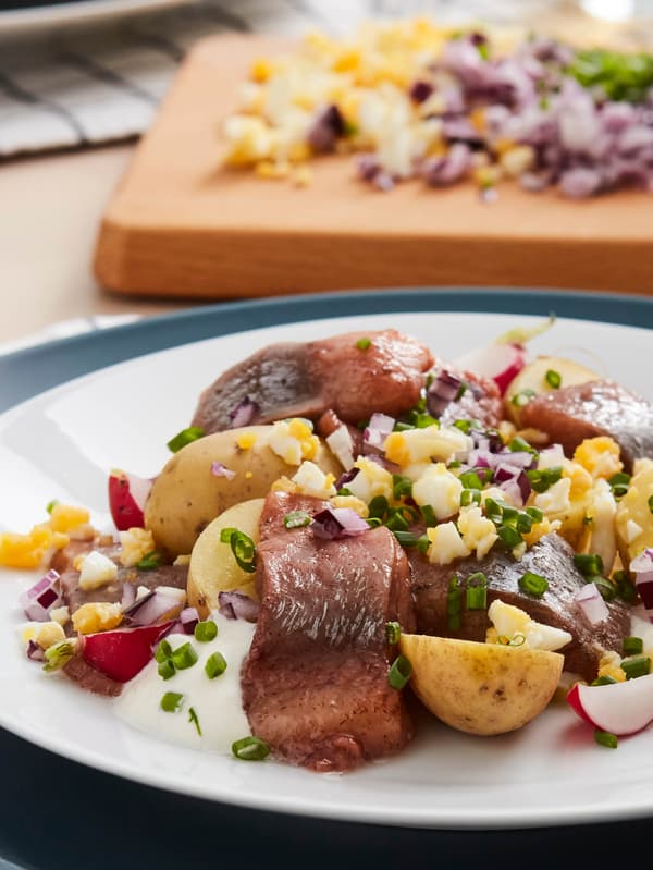 Šalát z nakladaných sleďov SILL MATJES so zemiakmi, reďkovkami, červenou cibuľou, vajíčkami a pažítkou na bielom tanieri s čiernou vidličkou TILLAGD.