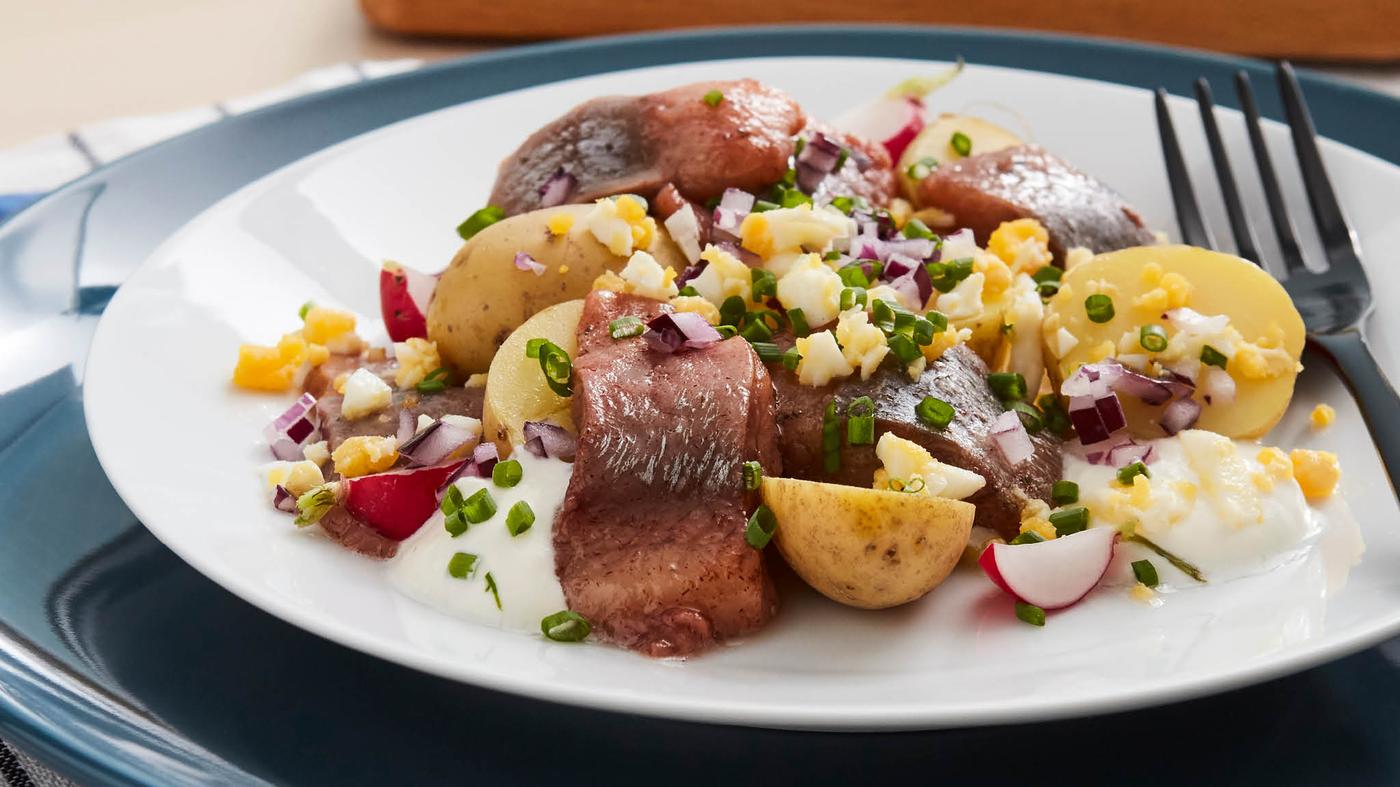 Šalát z nakladaných sleďov SILL MATJES so zemiakmi, s reďkovkami, červenou cibuľou, vajíčkami a pažítkou na bielom tanieri s čiernou vidličkou TILLAGD.