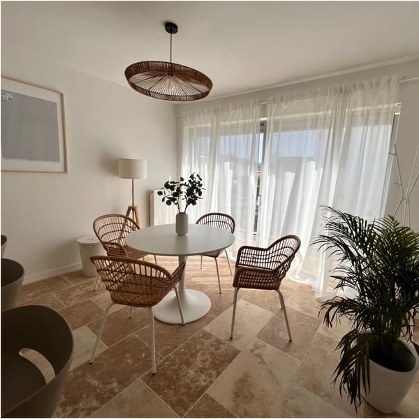 Salle à manger lumineuse avec table ronde blanche, chaises en rotin, rideaux transparents, plante verte et suspension naturelle au plafond.