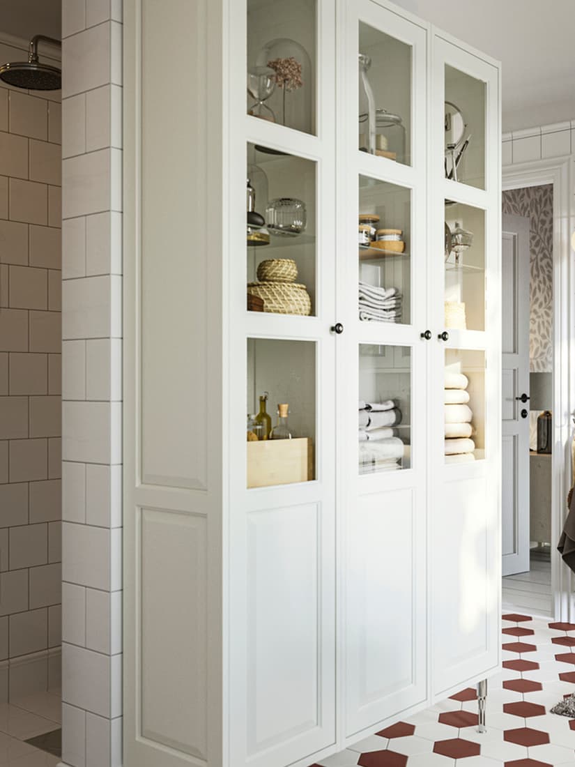 Salle de bain avec armoire colonne avec portes vitrées TÄNNFORSEN blanche remplie de serviettes, boîtes et flacons.