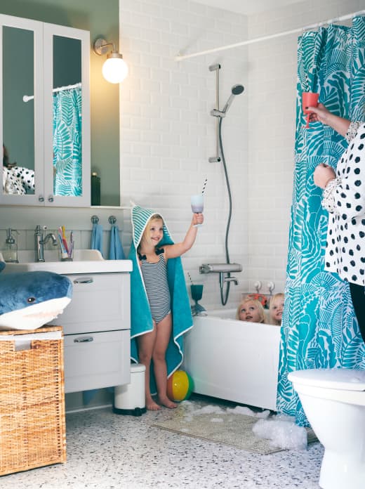 Salle de bain avec une baignoire et trois fillettes. L’une d’elle, enveloppée dans une serviette à capuche RÖRANDE, porte un toast imaginaire avec sa maman.