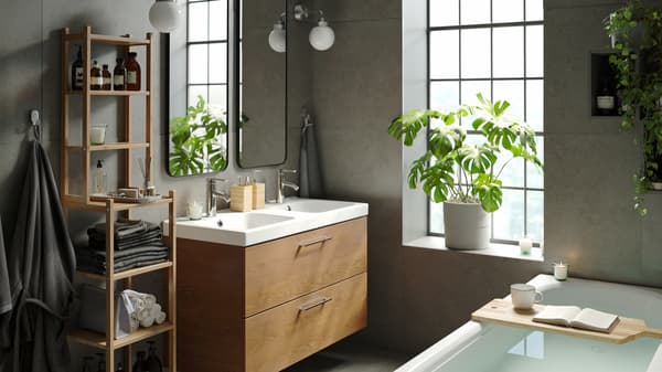 Salle de bain calme avec meuble lavabo teinté brun et étagère pour baignoire en bambou, deux miroirs, baignoire et plantes d’intérieure vertes.
