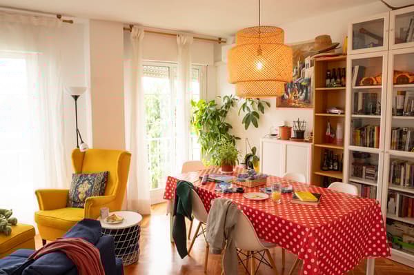 Salón antes de ser reformado con lámpara, estantería, mesa con hule, sillas, ropa, accesorios, sillón, ventana y cortinas.