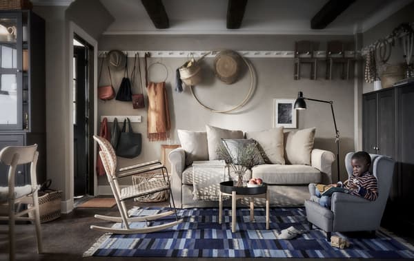 Salón con sofá, unha cadeira de brazos e un neno noutra pequena. Cuarto con estantes con tirador e almacenamento á vista.