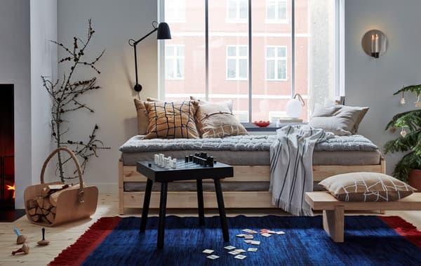 Salón dun apartamento con poucos mobles cunha cama encastelable baixo a fiestra, coxíns, téxtiles, dúas mesas auxiliares e unha alfombra grande.