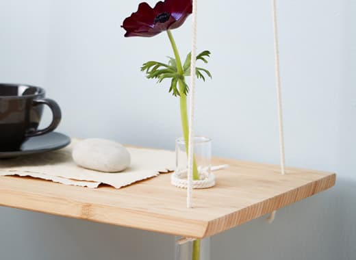 Samodzielnie zrobiony wiszący stolik nocny wykonany z deski do krojenia i liny z wywierconym otworem na długi szklany wazon. 