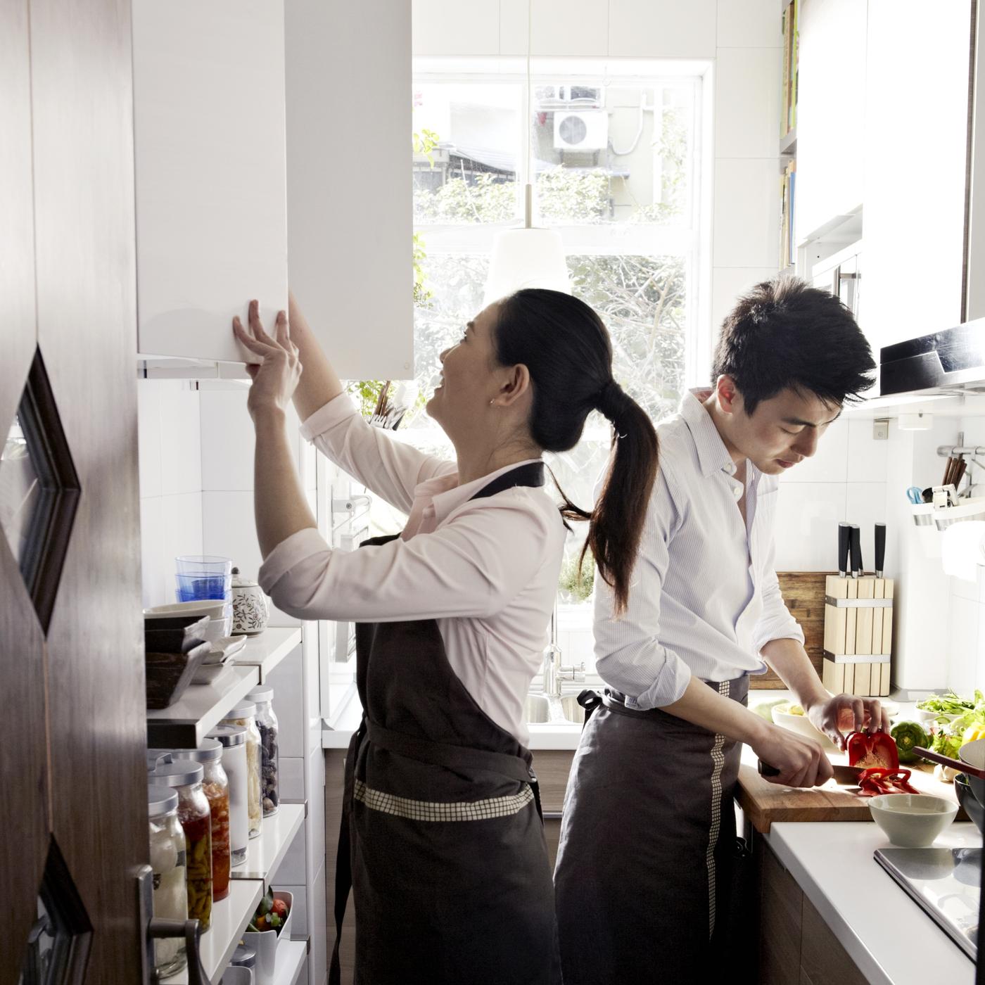 小さなキッチンスペースに、エプロンをつけた若者が2人いる。1人はキャビネットを開けていて、もう1人は野菜を刻んでいる。