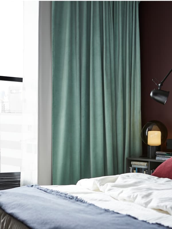 SANELA room darkening curtains in light green, a bed with a white quilt cover in a lightly lit bedroom.