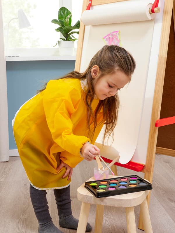 Sárga MÅLA hosszú ujjú kötényt viselő kislány, aki éppen MÅLA vízfestékkel fest.