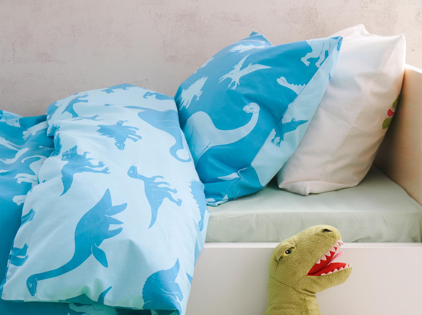 Sarung kuilt JÄTTELIK berwarna biru dengan cetakan dinosaur pada kedua-dua belah terletak di atas katil dengan mainan lembut dinosaur terjulur keluar dari bawah.