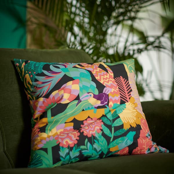 Sarung kusyen NÄBBFLY berwarna-warni yang menampilkan burung tropika di kalangan tumbuh-tumbuhan hijau yang subur dan pokok-pokok terletak di atas sofa baldu berwarna hijau.