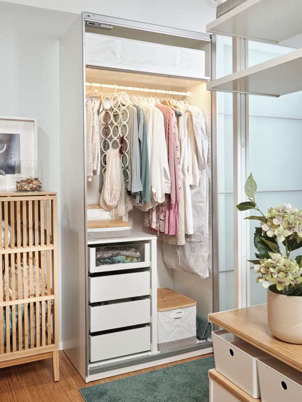 Šatníková skriňa PAX s posuvnými dverami NYKIRKE, vo vnútri sú štyri zásuvky, tri škatule RABBLA a veľa vešiakov s dámskym oblečením. Vľavo je časť bambusovej skrine NORDKISA. V popredí je vidieť časť úložnej zostavy ELVARLI.