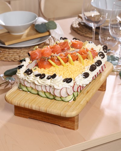 Savory sandwich cake decorated with smoked salmon, lemon slices, grated egg, cucumber, and radish on a wooden serving board, set on a festive dining table.