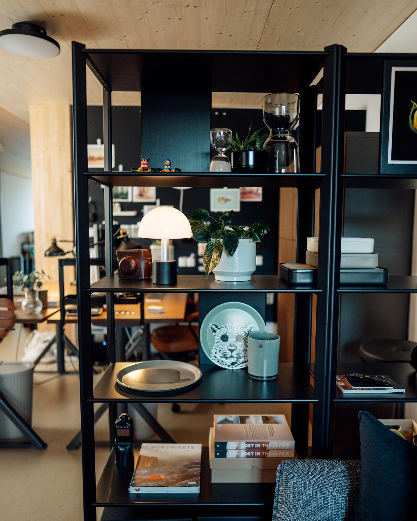 Scaffale nero aperto come divisorio, con libri, stoviglie, piante, lampada e accessori casa.
