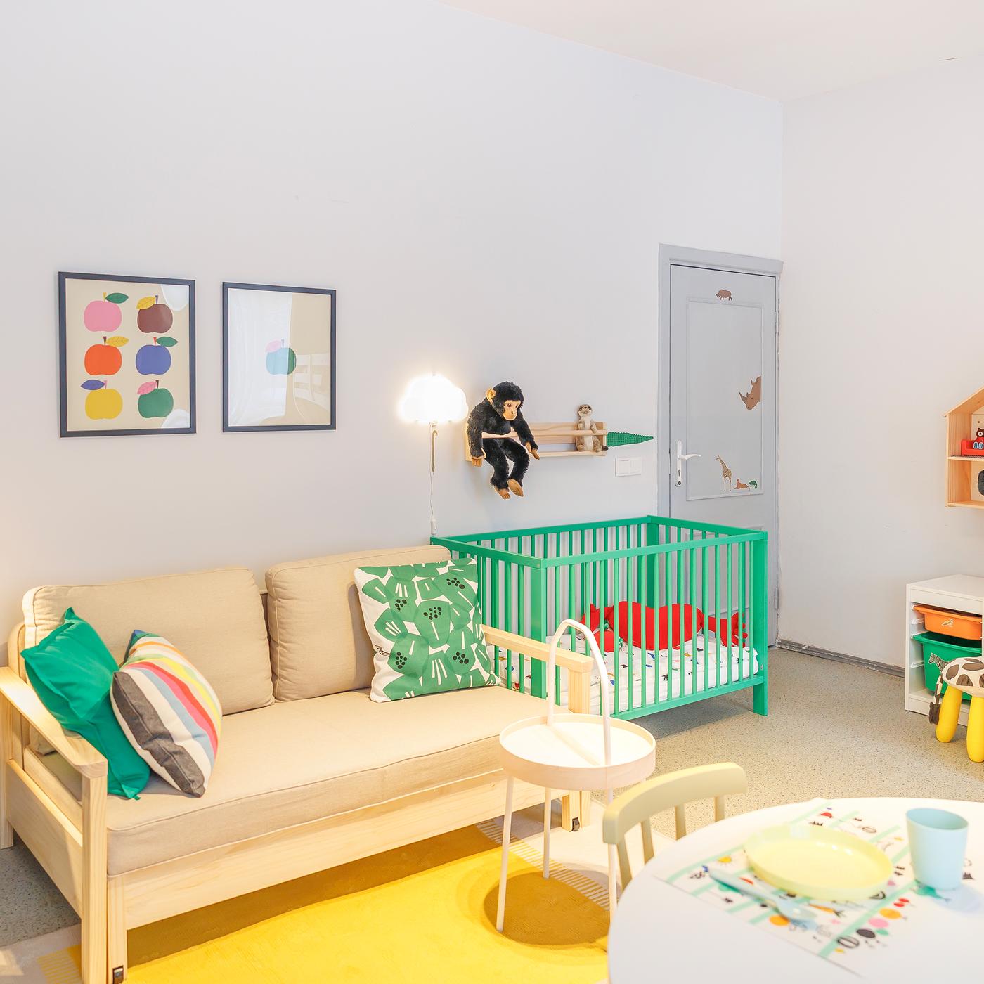 Scandinavian-style family room features a sofa bed, a round table with chairs and a green baby cot. The room is decorated with cushions, rugs, soft toys and wall art.