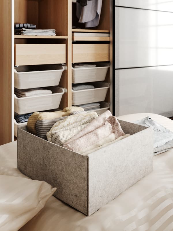 Scatola KOMPLEMENT su un letto, contenente indumenti ben piegati. Sullo sfondo, un guardaroba aperto, con vestiti appesi e cassetti.