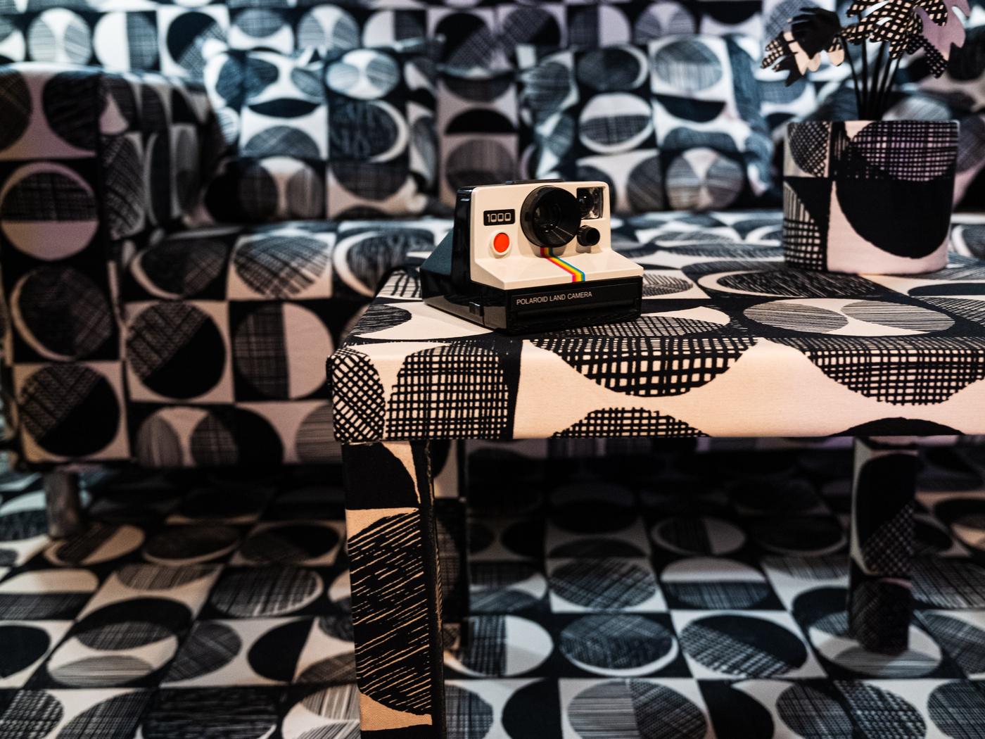 Schwarz weiß gefleckte Couch mit Couchtisch. Auf dem Tisch steht eine schwarz weiße Polaroid Kamera.  