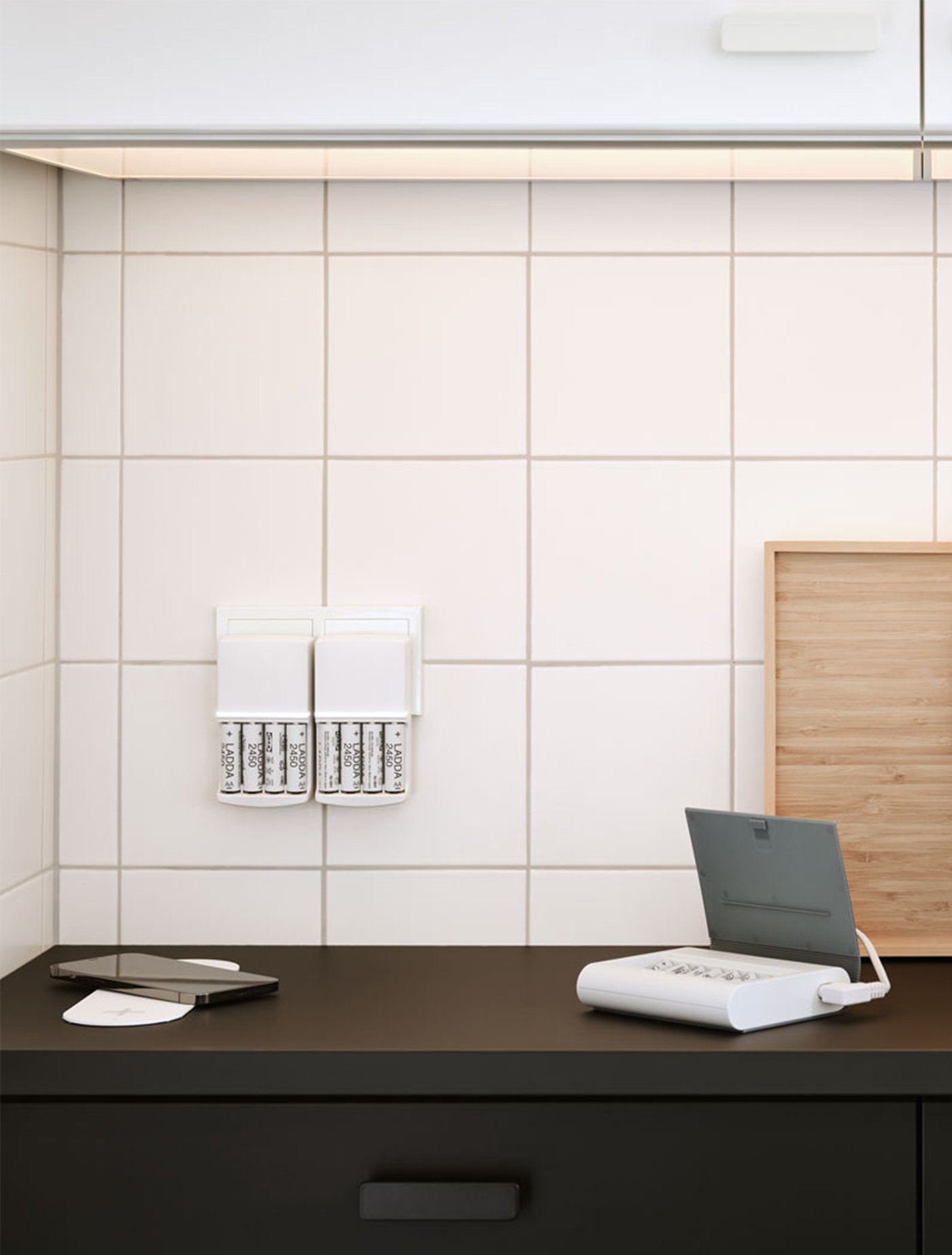 Schwarze Küche mit weißen Wandkacheln. Auf der Arbeitsplatte liegt ein Ladegerät für ein Smartphone, ein Akku und eine Batterieladestation an der Wand.