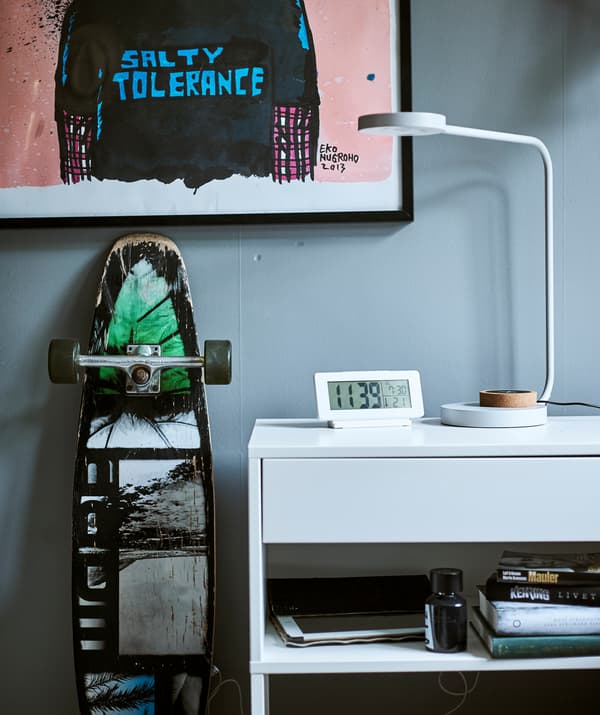 Scrivania bianca con ripiano integrato, lampada da tavolo e orologio digitale, uno skateboard e una stampa in stile graffiti.