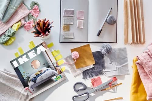 Scrivania da lavoro creativa con campioni di tessuto colorati, gomitoli, matite e forbici intorno a un quaderno aperto e al catalogo IKEA. 