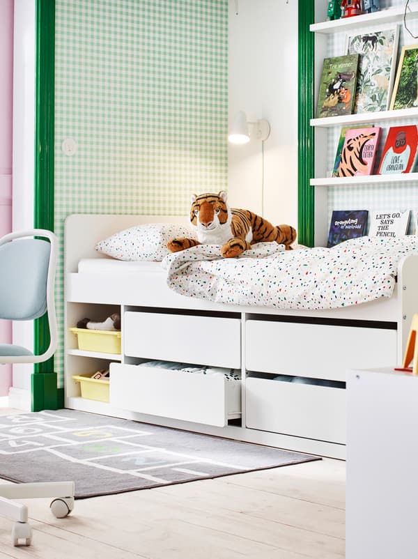 Secção de um quarto de criança em verde e branco, com livros em prateleiras para molduras, brinquedos e uma estrutura de cama SLÄKT com arrumação em baixo.