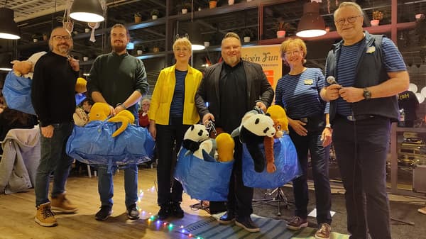 Sechs lächelnde Menschen stehen im IKEA Restaurant, ein IKEA Mitarbeiter, zwei IKEA Mitarbeiterinnen, jeweils in IKEA Bekleidung in blau und gelb. Die anderen Männer sind dunkel gekleidet. Zwei tragen prall gefüllte blaue FRAKTA Taschen, die gefüllt sind mit Stofftieren. Pandabären, ein Affe, ein Hai, eine Schlange und eine Krake schauen aus den Taschen raus. Auf dem Boden liegt eine bunte Lichterkette. 