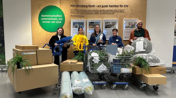 Sechs Personen stehen in einem IKEA-Markt vor einer Holztafel mit der Aufschrift „Gemeinschaften unterstützen, wo immer wir sind“. Sie sind umgeben von Einkaufswagen und Transportwagen voller Möbel, Pflanzen, Kissen und Haushaltswaren. Zwei der Personen tragen IKEA-Mitarbeiterkleidung, während die anderen in Freizeitkleidung gekleidet sind.  