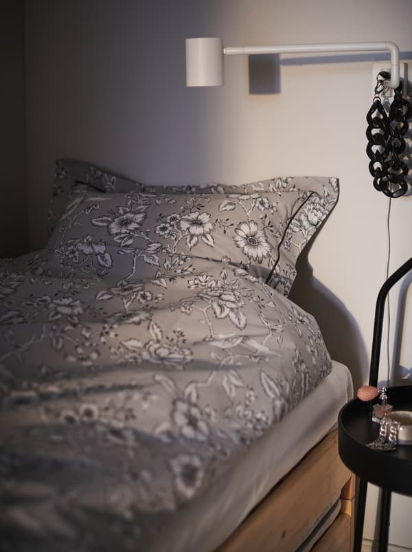 Section of a bed made with floral-patterned PRAKTBRÄCKA bed linen. A NYMÅNE wall lamp is poised for bedtime reading.