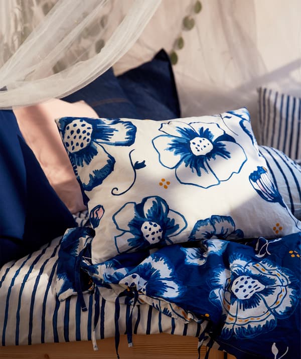 Section of a bed with bed linen, pillows and cushions in a blue-white mix of floral patterns, pinstripes and unicolour.