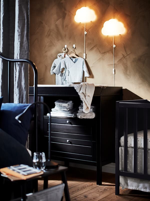 Section of a bedroom, half hidden behind a room-dividing curtain, with a black-brown SUNDVIK changing table and a baby’s crib.