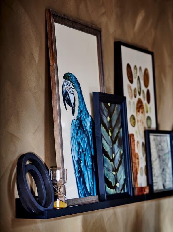Section of a brown wall with a black MOSSLANDA picture ledge holding various artful objects and framed pictures.