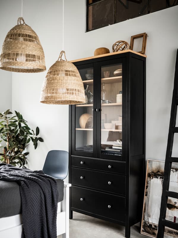 Section of a light bedroom with a tall, black HEMNES glass-door cabinet, TORARED pendant lamps, decorations and a plant.