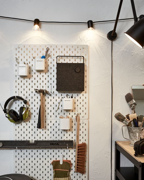 Section of a wall fitted with a pegboard holding tools, earphones and a bluetooth speaker; lighting chain and lamp.