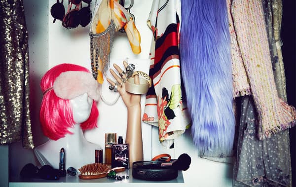 Section of a wardrobe filled with flamboyant party clothing and accessories; bust with neon wig, mannequin hand with rings.
