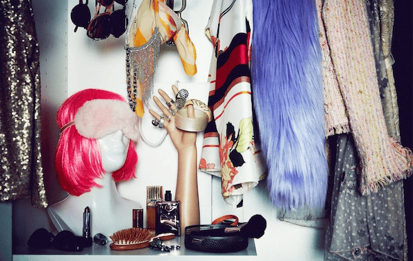 Section of a wardrobe filled with flamboyant party clothing and accessories; bust with neon wig, mannequin hand with rings.