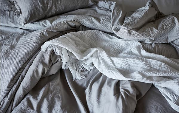 Section of an empty, slept-in bed with creased, fluffy, one-colour bedlinen; a throw untidily lying on top.