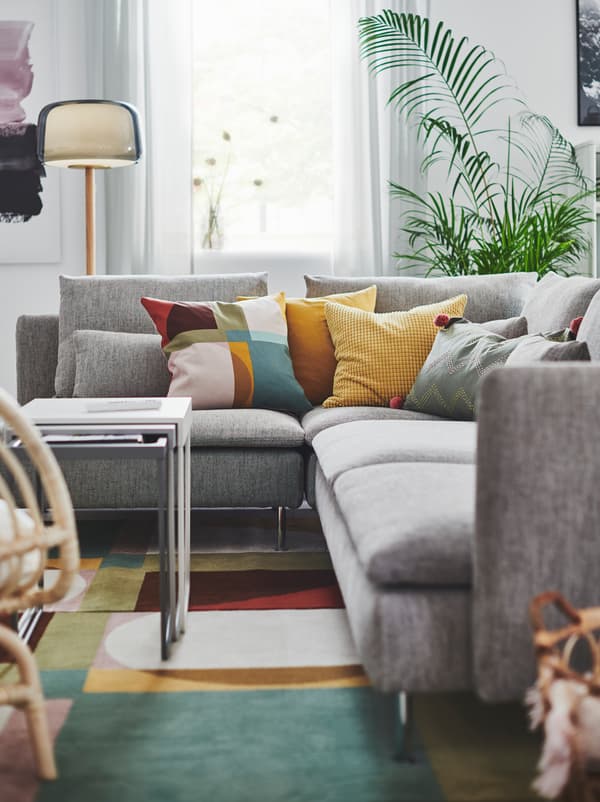Section of living room with a grey SÖDERHAMN corner sofa, a host of colourful cushions on it and standing on a colourful rug.