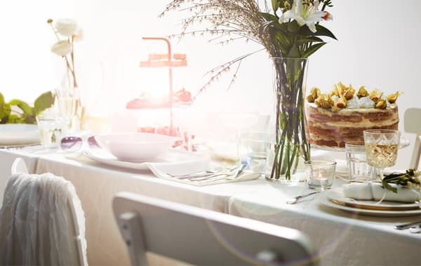 Section of long table set in a romantic, lavish way with white furniture, cloth and tableware; cake, cookie tray and flowers.
