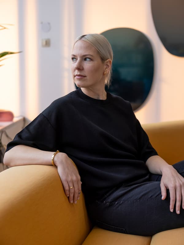 Seduta su un divano giallo nel suo studio, la designer Sabine Marcelis, vestita di nero, rivolge lo sguardo alla sua destra.