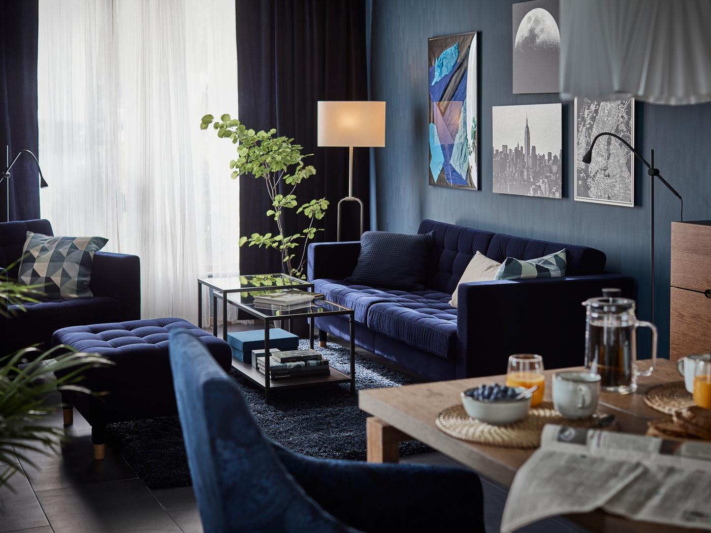 Séjour avec œuvres d’art, plantes vertes et canapé, repose-pieds et fauteuil LANDSKRONA en bleu foncé, autour de tables gigognes VITTSJÖ.