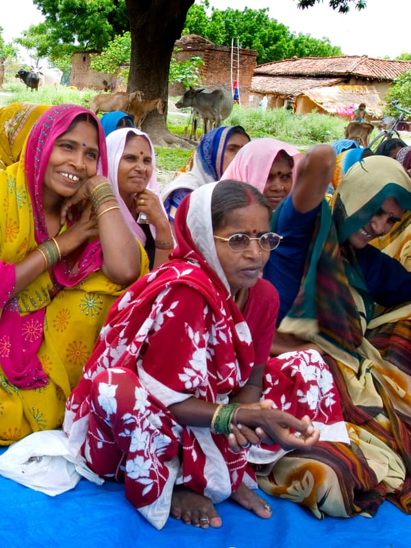 Sekumpulan wanita India yang memakai pakaian berwarna terang sedang duduk di atas ambal di ruang luar. Beberapa pondok dan lembu kelihatan di belakang.