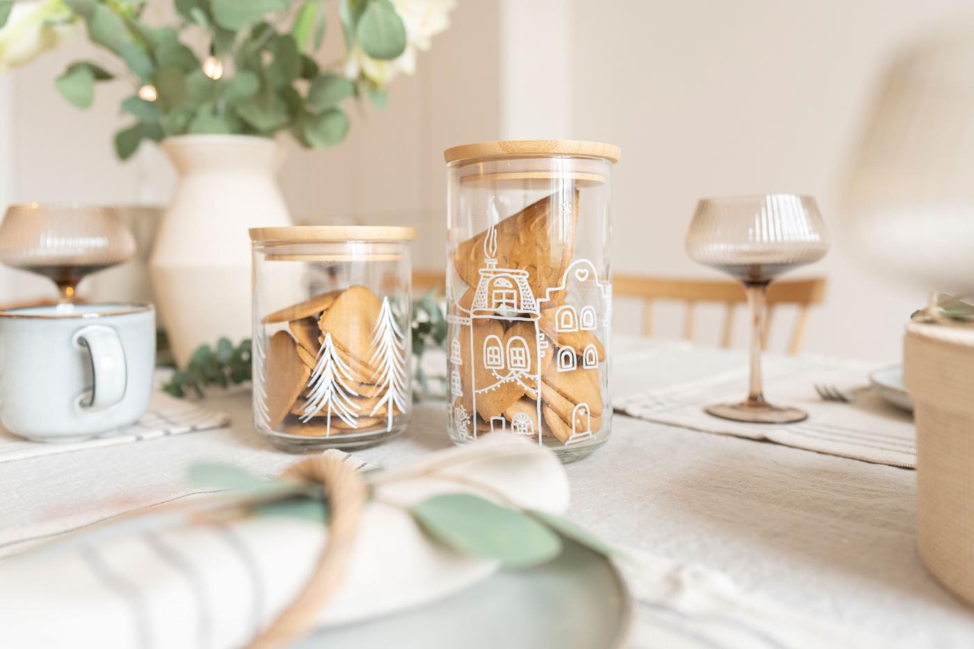 Selbstbemalte Vorratsgläser EKLATANT gefüllt mit Weihnachtsplätzchen auf einem gedeckten Tisch. 