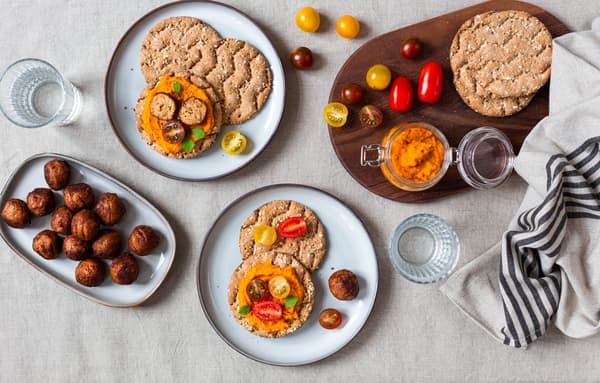 Selbstgemachter Kürbis-Karotten-Aufstrich steht in einem Glas angerichtet auf einem Tisch mit Knäckebrot und Proteinbällchen.