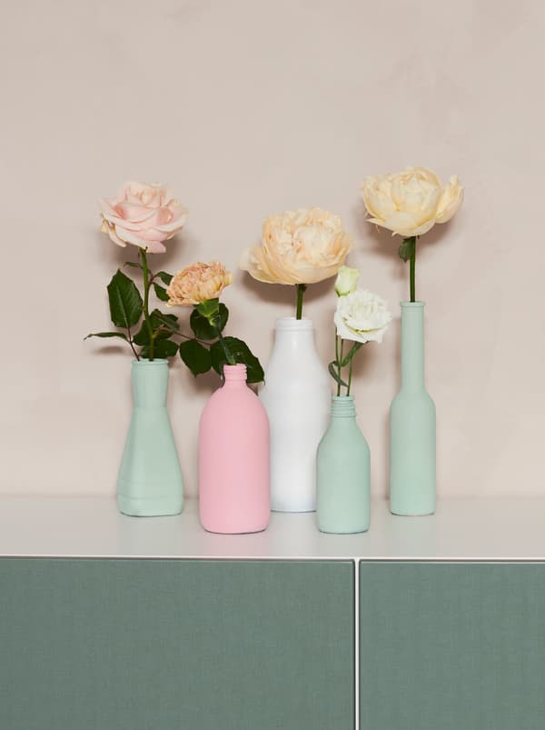 Selonggok botol lama pelbagai bentuk yang telah dicat dengan warna pastel dan dijadikan vas, setiap satu mempunyai bunga SMYCKA.