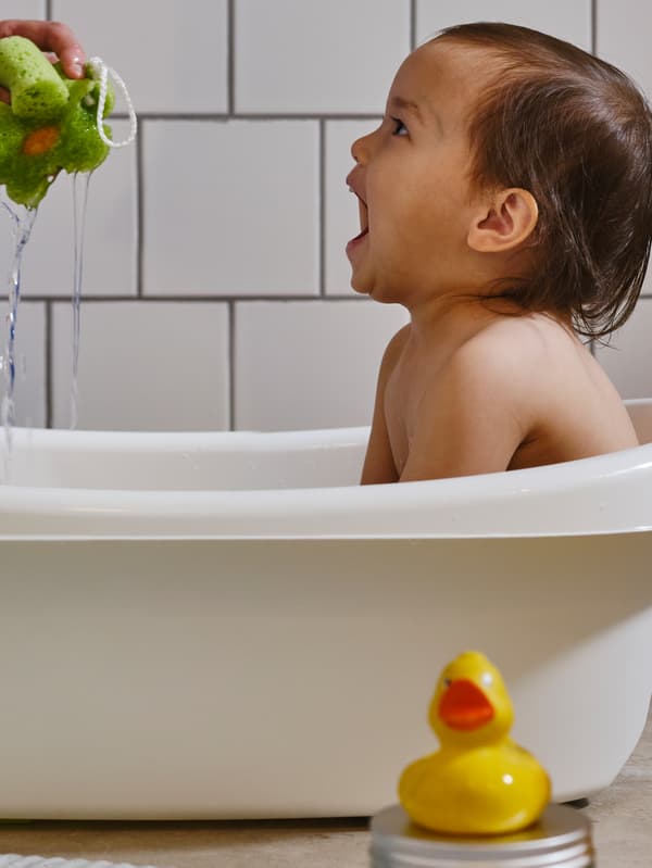 Seorang budak kecil yang duduk di dalam tab mandi LÄTTSAM berwarna putih sedang ketawa sementara tangan seorang dewasa memerah span berwarna hijau.