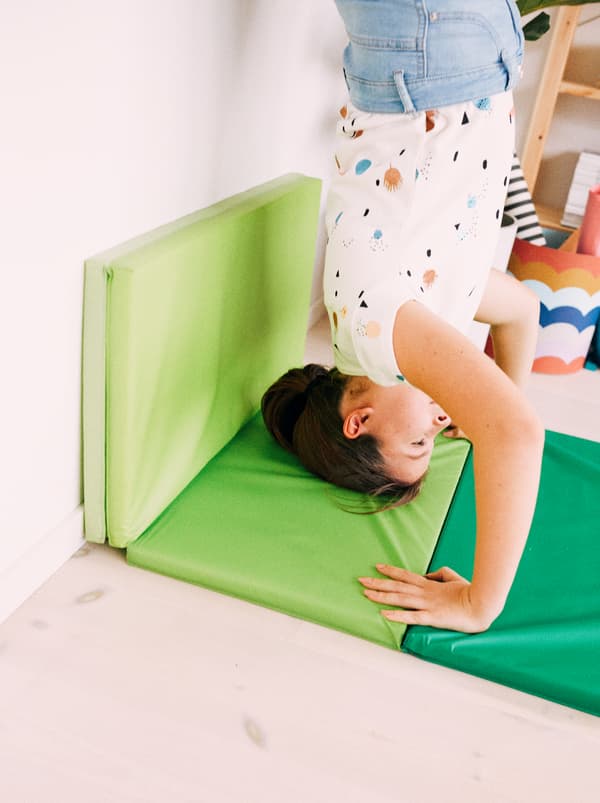 Seorang budak perempuan melakukan dirian tangan dengan bersandar pada dinding, kepala dan tangannya disokong oleh tikar gim lipat PLUFSIG berwarna hijau terang.