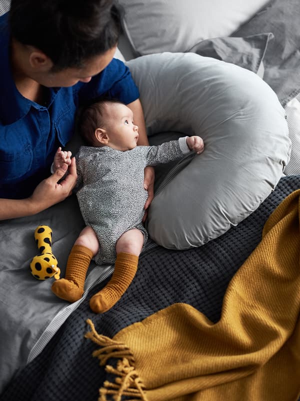 Seorang ibu dan bayinya duduk di atas katil dengan sarung berwarna kelabu dan mustard, dan bantal susuan di sisi mereka.
